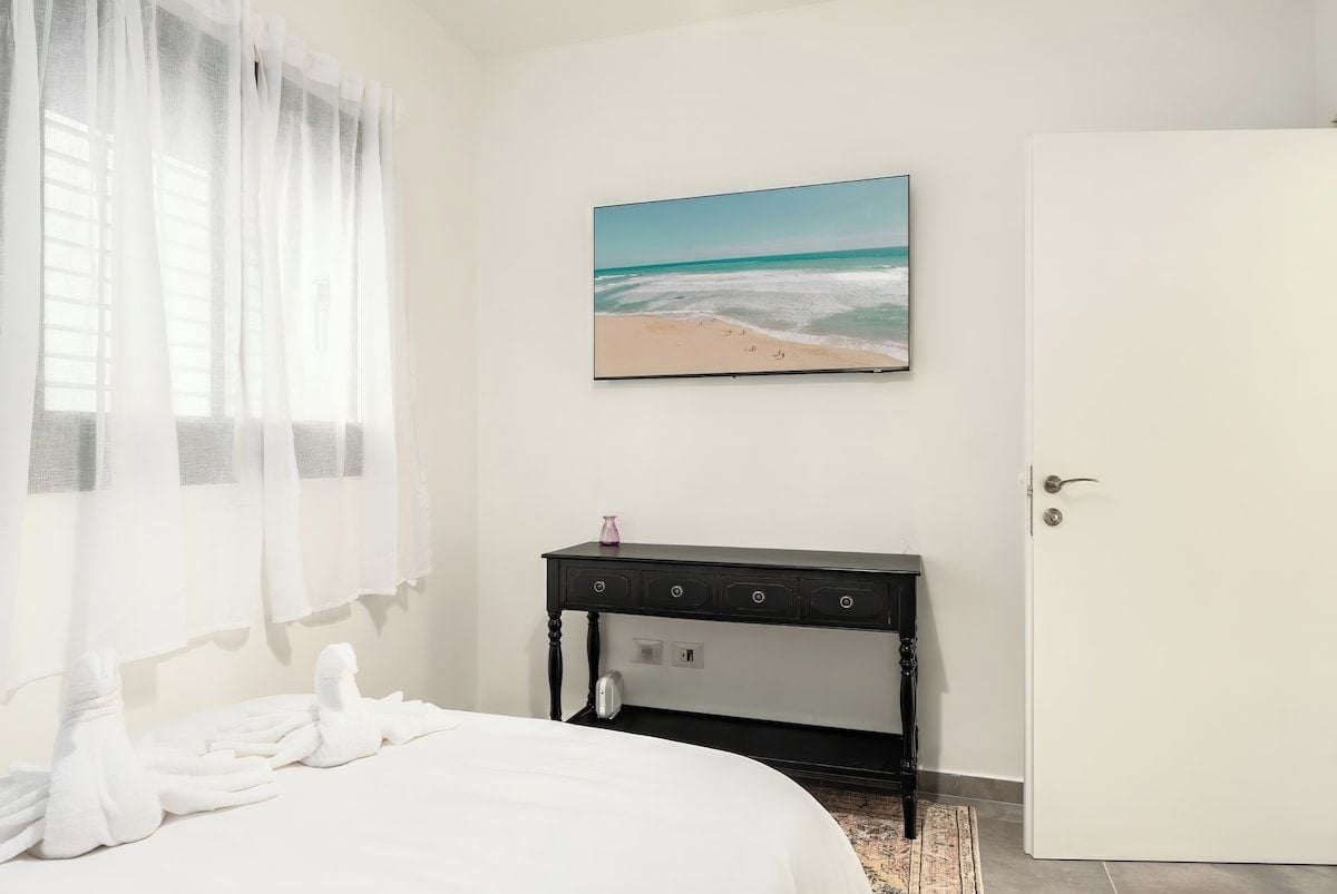 A hotel room with a bed, a desk, a painting of a beach, and a door.