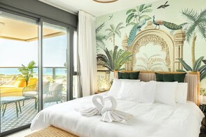 Une chambre d’hôtel avec un grand lit, un balcon avec des chaises jaunes et une fresque représentant des oiseaux et des plantes.