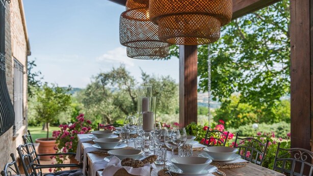 Un espace de restauration en plein air avec une table en bois dressée pour un repas, entourée de chaises et d’une pergola agrémentée de guirlandes lumineuses.