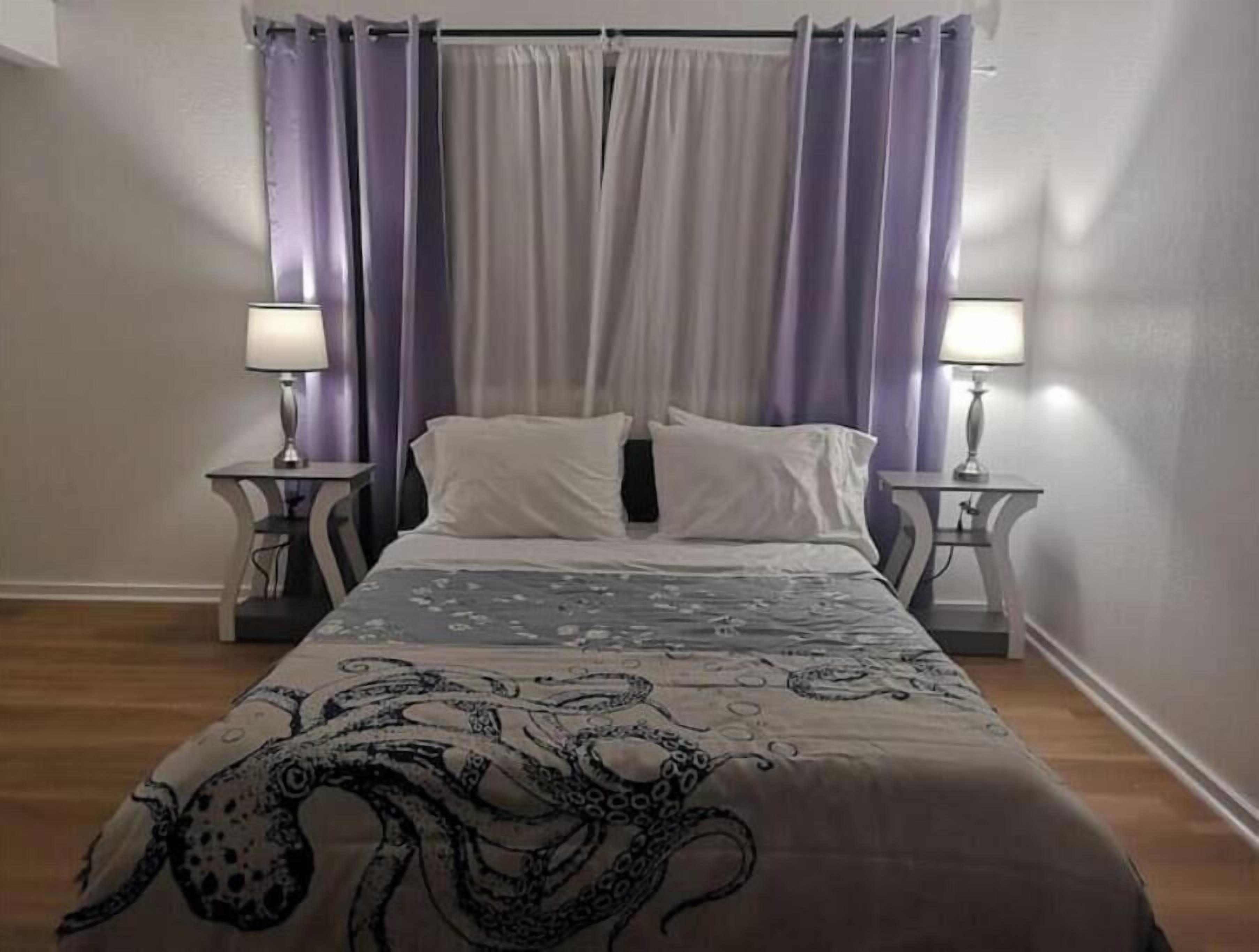 Un dormitorio con una cama, dos mesitas de noche y un ventanal con cortinas moradas.