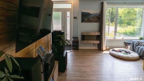 A modern living room with a large flat-screen TV, wooden flooring, a fireplace, and a view of greenery through large windows. - LITTLE PARADISE IN NEUVILLE, NATURE AND TRANQUILITY (Neuville)