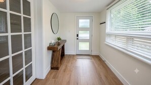 A hallway with a wooden floor, a white door with frosted glass panels, a small wooden console table with a plant, and a large window with blinds. - Renovated Ravine Cottage | Walk to Beach | Sleeps 8 | A/C, BBQ, & Firepit (Port Stanley)