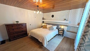 A bedroom with a bed, dresser, nightstands, and a brick accent wall. - Renovated Ravine Cottage | Walk to Beach | Sleeps 8 | A/C, BBQ, & Firepit (Port Stanley)
