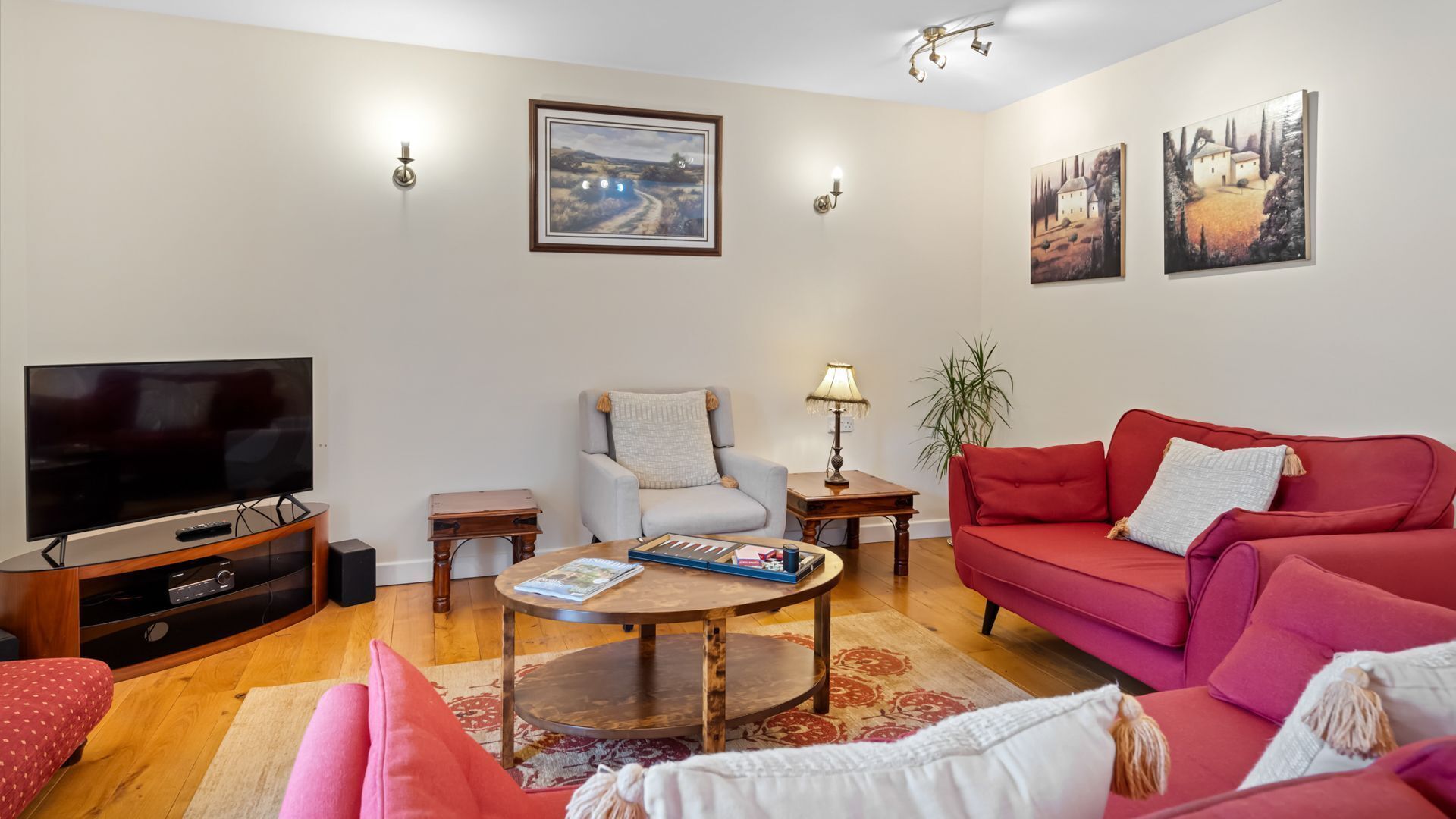 A living room with a flat-screen TV, a red sofa, a grey armchair, a wooden coffee table, and artwork on the walls.