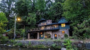 Ein zweistöckiges Steinhaus mit Balkon und steinernen Hauseingang, vor dem ein Fluss fließt.