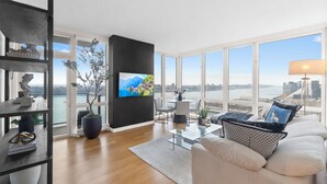 Un saló modern amb un televisor gran, un sofà, una taula de centre i vista a un paisatge urbà a través de finestres grans.