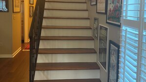 Una scala con gradini in legno e una ringhiera nera, che conduce a un pianerottolo con una porta. Le pareti sono decorate con quadri e opere d'arte.