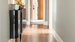 Un pasillo con piso de madera, una consola negra con base dorada y un espejo con lámpara blanca.