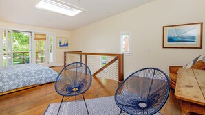 A spacious bedroom with a large bed, two blue wire chairs, a wooden table, and a view of greenery through the windows. - Charming Sag Harbor Apartment. Two blocks from Main and Bay Streets. (Sag Harbor)
