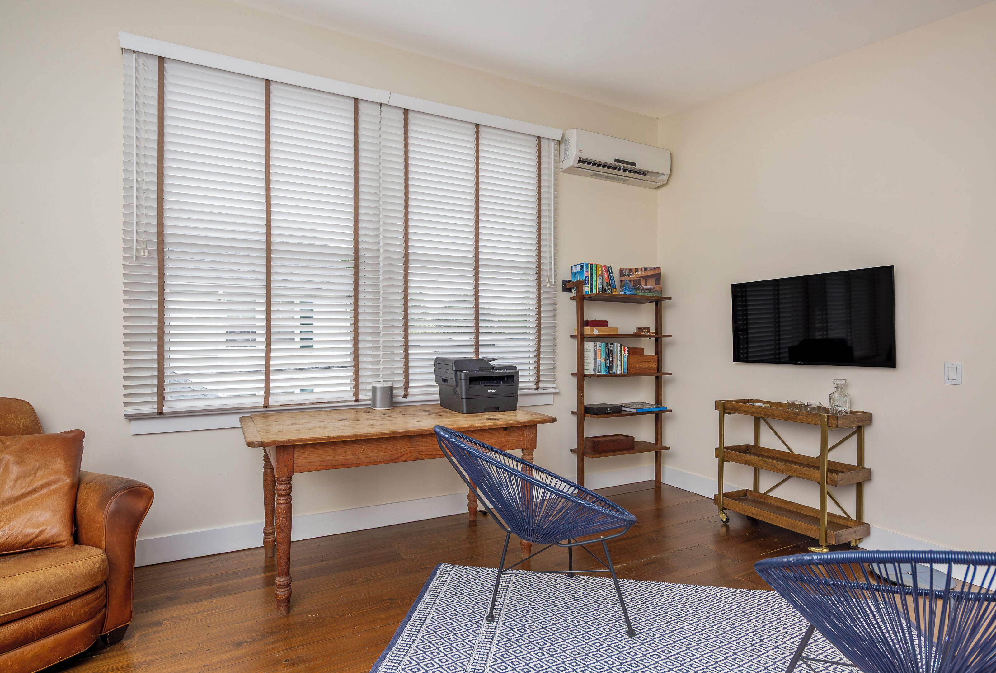 A living room with a wooden desk, a blue chair, a leather couch, a bookshelf, and a mounted TV.