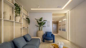 A modern living room with a grey sofa, a blue armchair, and a wooden coffee table. There are shelves with plants and decorative items, and a large potted plant in the center. - Apart With Barbecue 600m From the Beach Aqt0604 (Itapema)