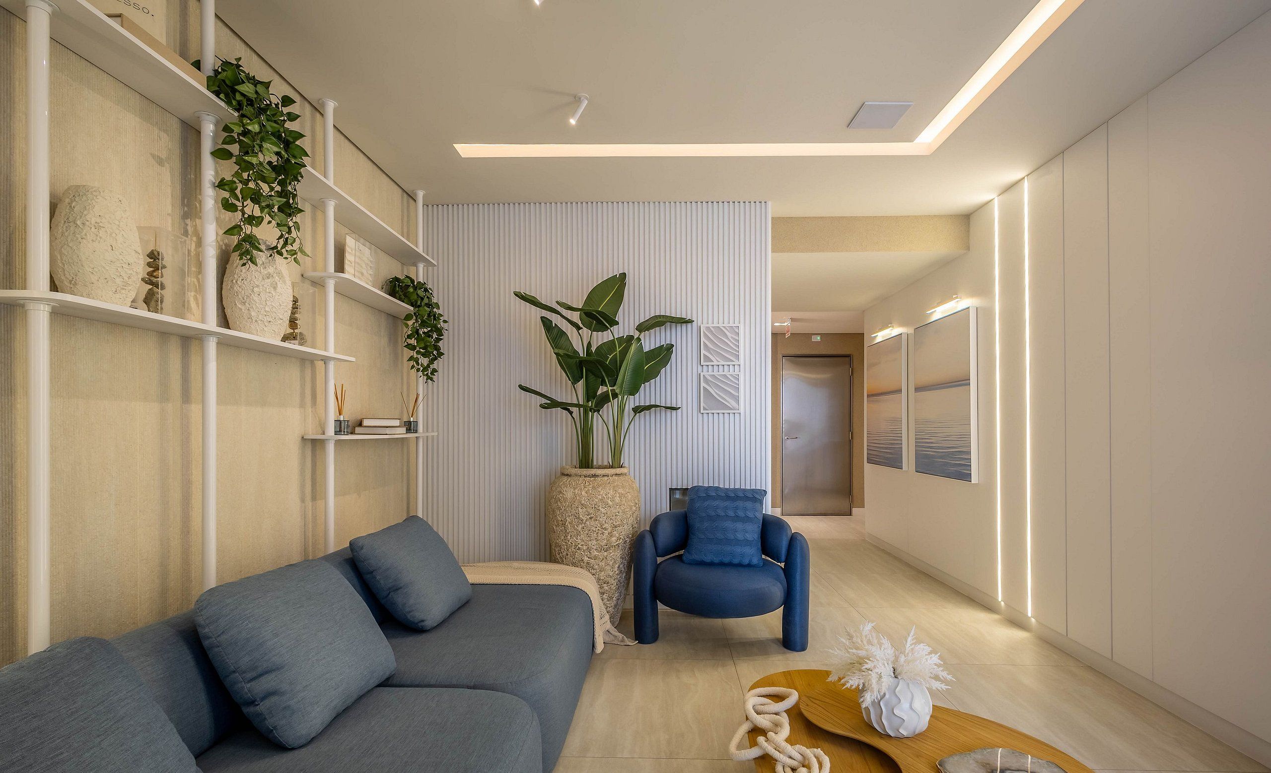 Ein modernes Wohnzimmer mit einer grauen Couch, einem blauen Sessel und einem Holztisch. Es gibt Regale mit Pflanzen und Dekorationsgegenständen sowie eine große Pflanze in der Mitte.
