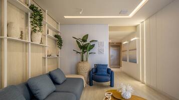 Un salón moderno con un sofá gris, un sillón azul y una mesa de centro de madera. Hay estanterías con plantas y objetos decorativos, y una planta grande en maceta en el centro.