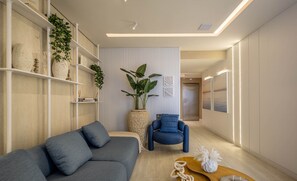 A modern living room with a grey sofa, a blue armchair, and a wooden coffee table. There are shelves with plants and decorative items, and a large potted plant in the center.