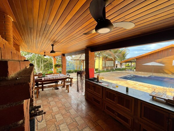 A covered outdoor kitchen with a brick oven, wooden table, and bar area overlooking a swimming pool.