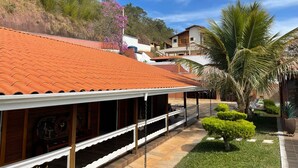 A building with a red-tiled roof, a covered outdoor area with a white railing, and a well-maintained garden with palm trees and shrubs. - Beautiful farm in Igarata right on the dam. (Canto das Águas Igaratá)