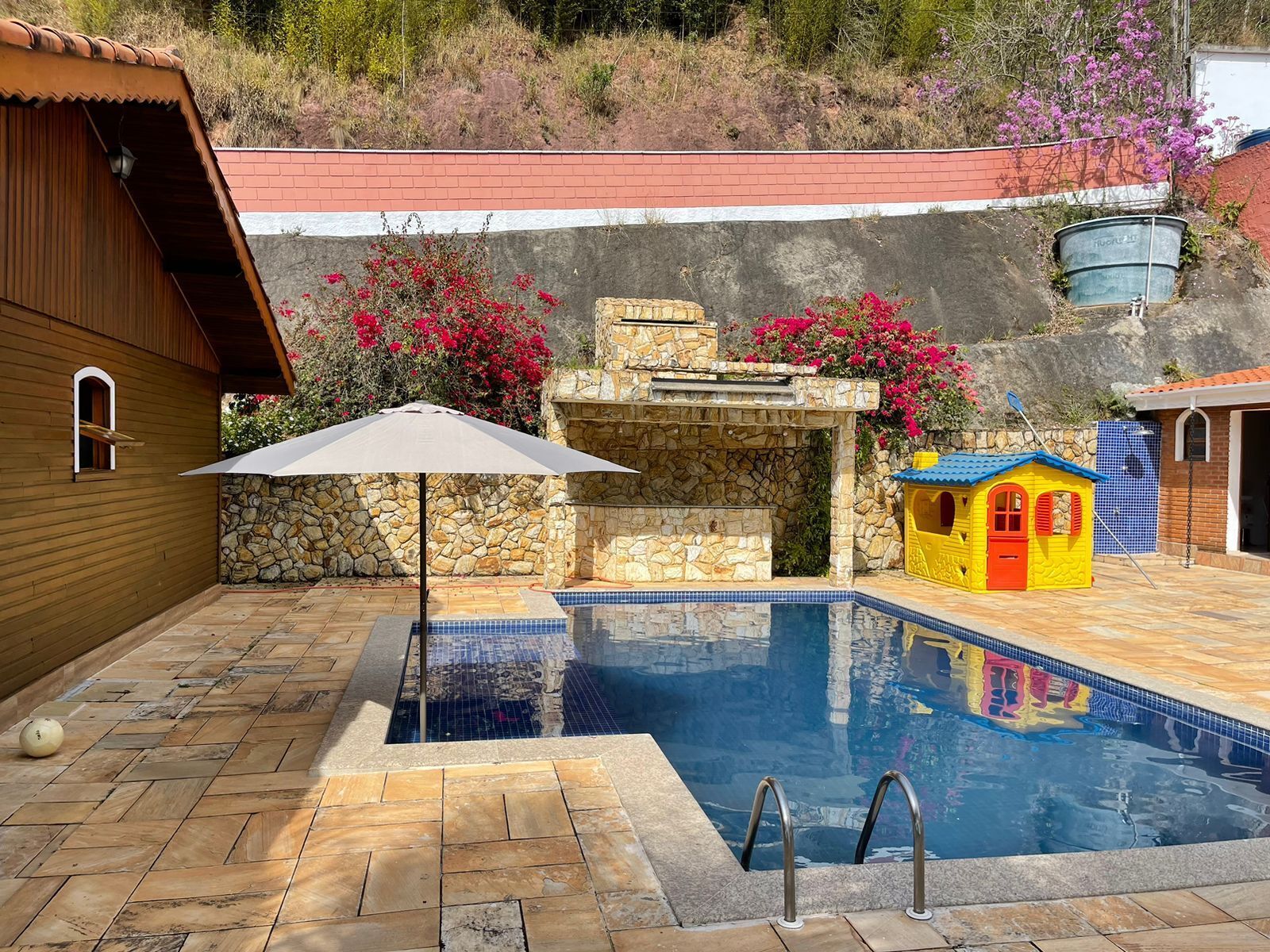 Piscina com paredes de pedra, uma casa pequena e um edifício com telhado vermelho.