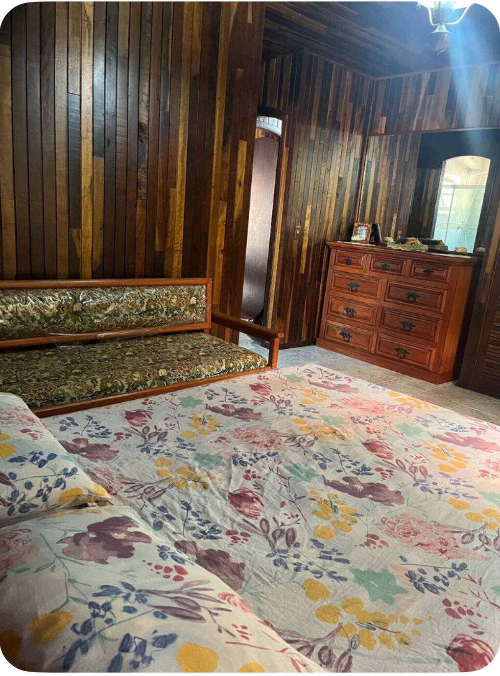 A room with a wooden wall panel, a floral-patterned bedspread, a bench with a floral cushion, and a wooden dresser with drawers.