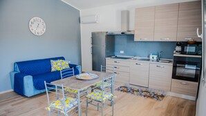 A compact living space with a kitchenette, a dining area with a table and chairs, a blue sofa, and a clock on the wall. - Villa Rosy - Your Villa Near the Sea (Marina di Butera)