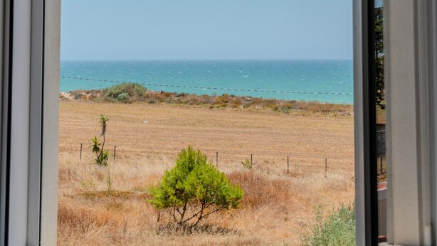 Blick durch ein Fenster auf eine Küstenlandschaft mit blauem Himmel, einem Sandstrand und spärlicher Vegetation.
