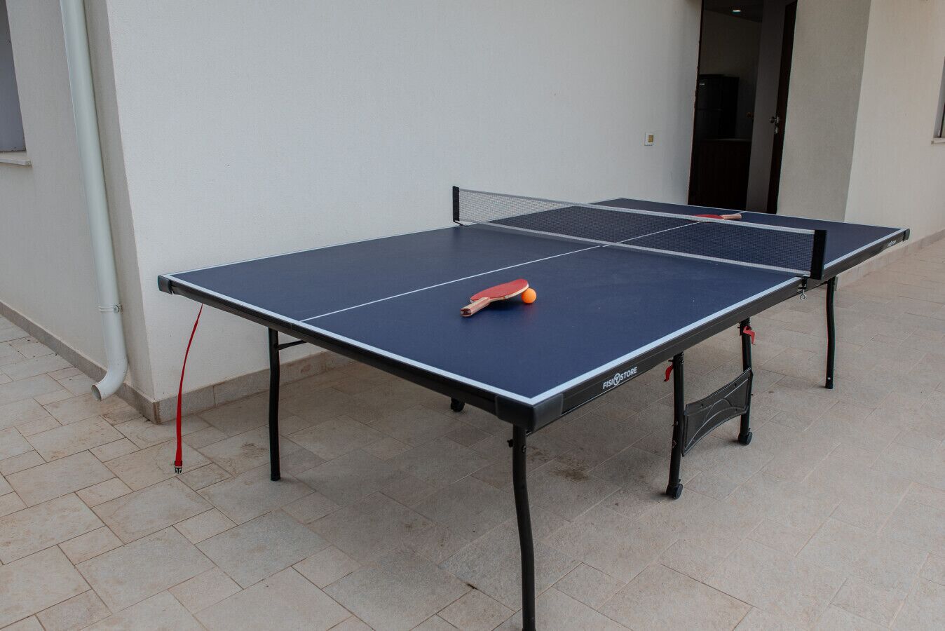 A ping pong table with a paddle and ball, set up outdoors on a tiled floor.