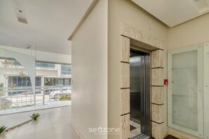 Een modern gebouw met een glazen lift, een balkon met beplanting en uitzicht op de straat.