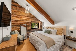 A bedroom with a wooden log wall, a bed with multiple pillows, a bedside table, a lamp, and a window with a view.