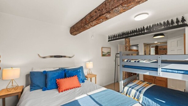 A bedroom with a bunk bed, a bed with blue bedding, a nightstand with a lamp, and a rustic ceiling beam.