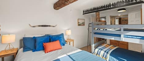 A bedroom with a bunk bed, a bed with blue bedding, a nightstand with a lamp, and a rustic ceiling beam.