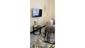 Een woonkamer met een flatscreen televisie aan de muur, een bijzettafeltje met een boek en een afstandsbediening, een ronde salontafel en een beige bank met een kussen.