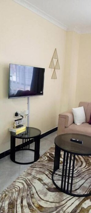 A living room with a flat-screen TV mounted on the wall, a small side table with a book and a remote, a round coffee table, and a beige sofa with a cushion. - Sara Spaces (Kiambu)