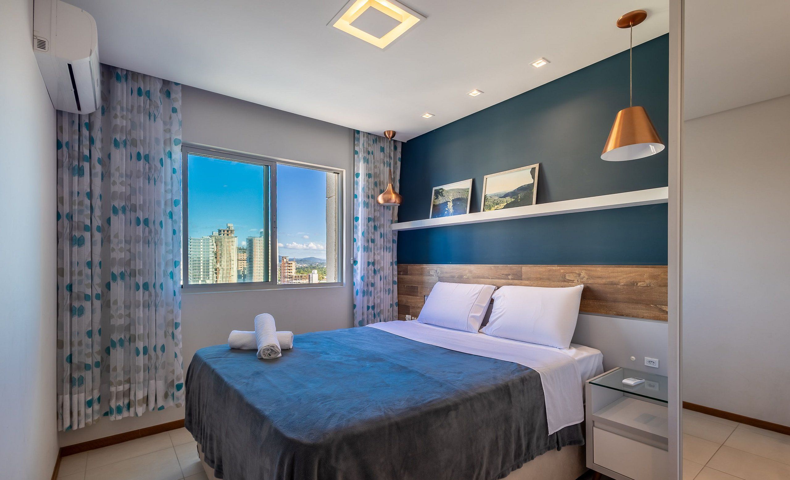 Habitación de hotel con cama, mesita de noche, repisa en la pared y ventana con vista a la ciudad.
