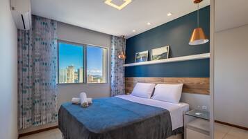 Habitación de hotel con cama, mesita de noche, repisa en la pared y ventana con vista a la ciudad.