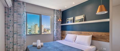 A hotel room with a bed, a nightstand, a lamp, a window with a city view, and an air conditioning unit.