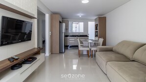 Una sala de estar moderna con una televisión de pantalla plana, un sofá, un comedor con mesa y sillas, y una cocina al fondo.