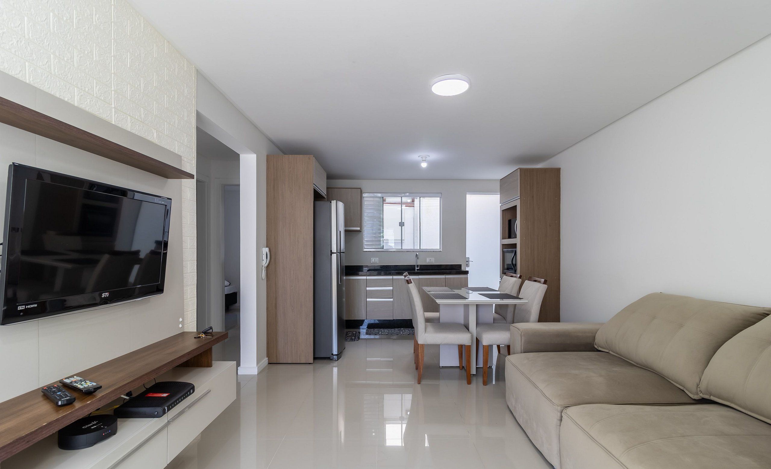 A modern living room with a flat-screen TV, a sofa, a dining area with a table and chairs, and a kitchen in the background.