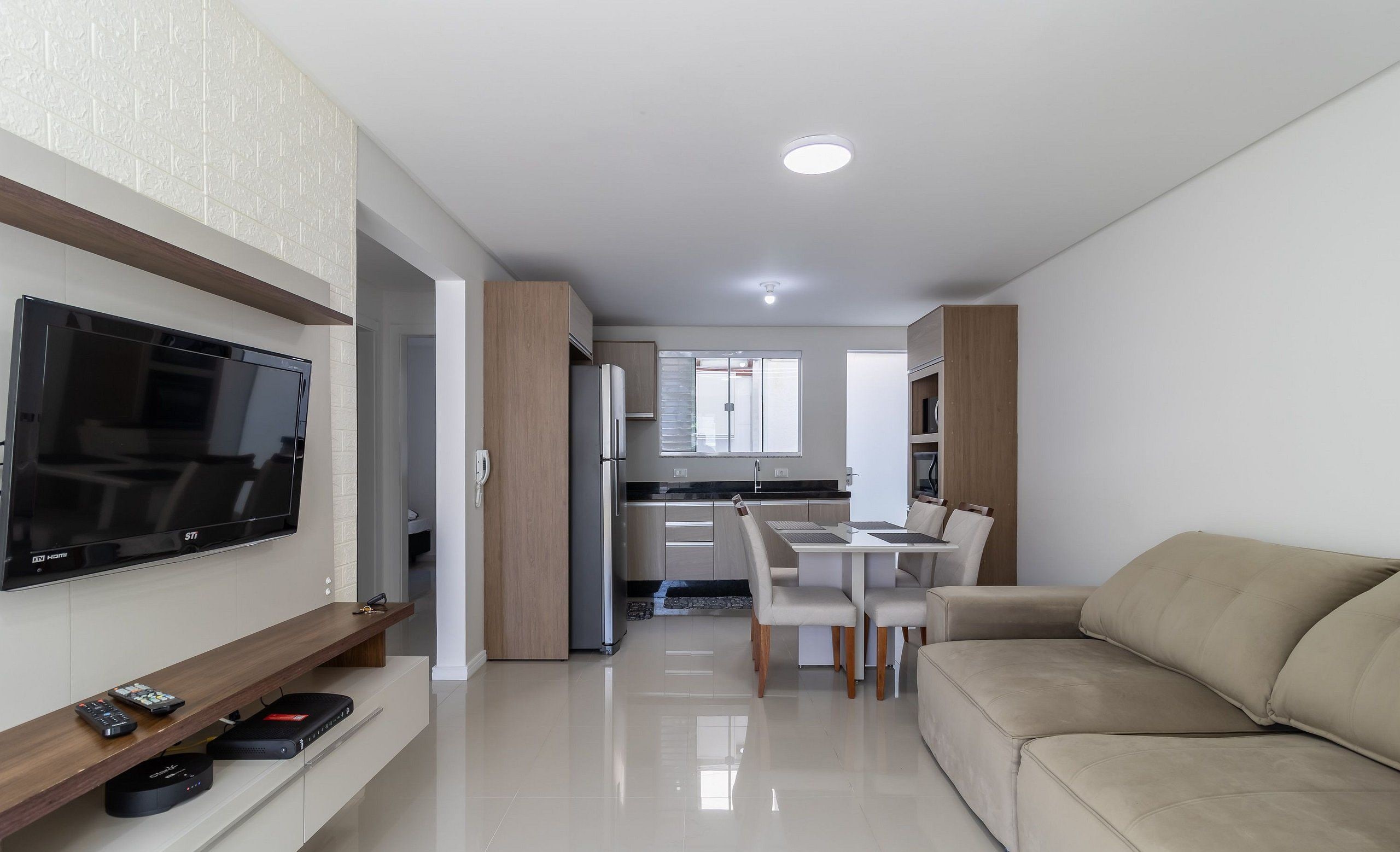 A modern living room with a flat-screen TV, a sofa, a dining area with a table and chairs, and a kitchen in the background.