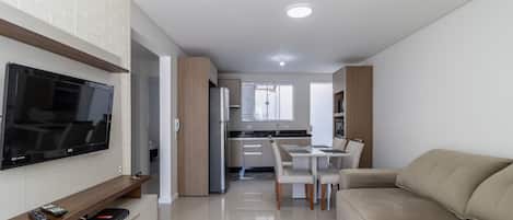 Una sala de estar moderna con una televisión de pantalla plana, un sofá, un comedor con mesa y sillas, y una cocina al fondo.