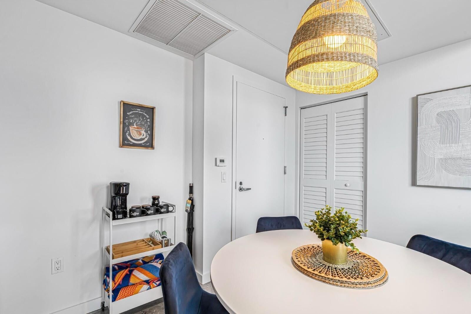 A modern, well-lit room with a round table, a hanging wicker light fixture, a coffee station, and a framed picture on the wall.