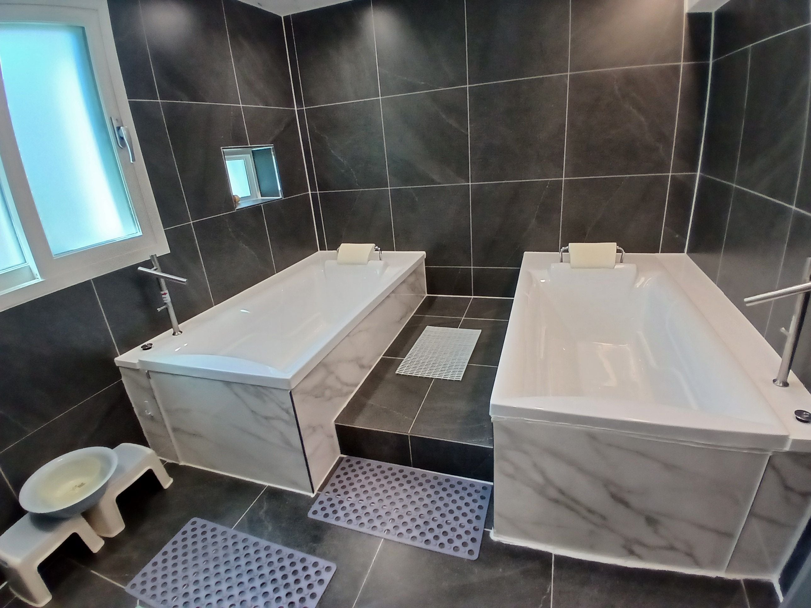 A modern bathroom with two separate tubs, a small stool, and a window.