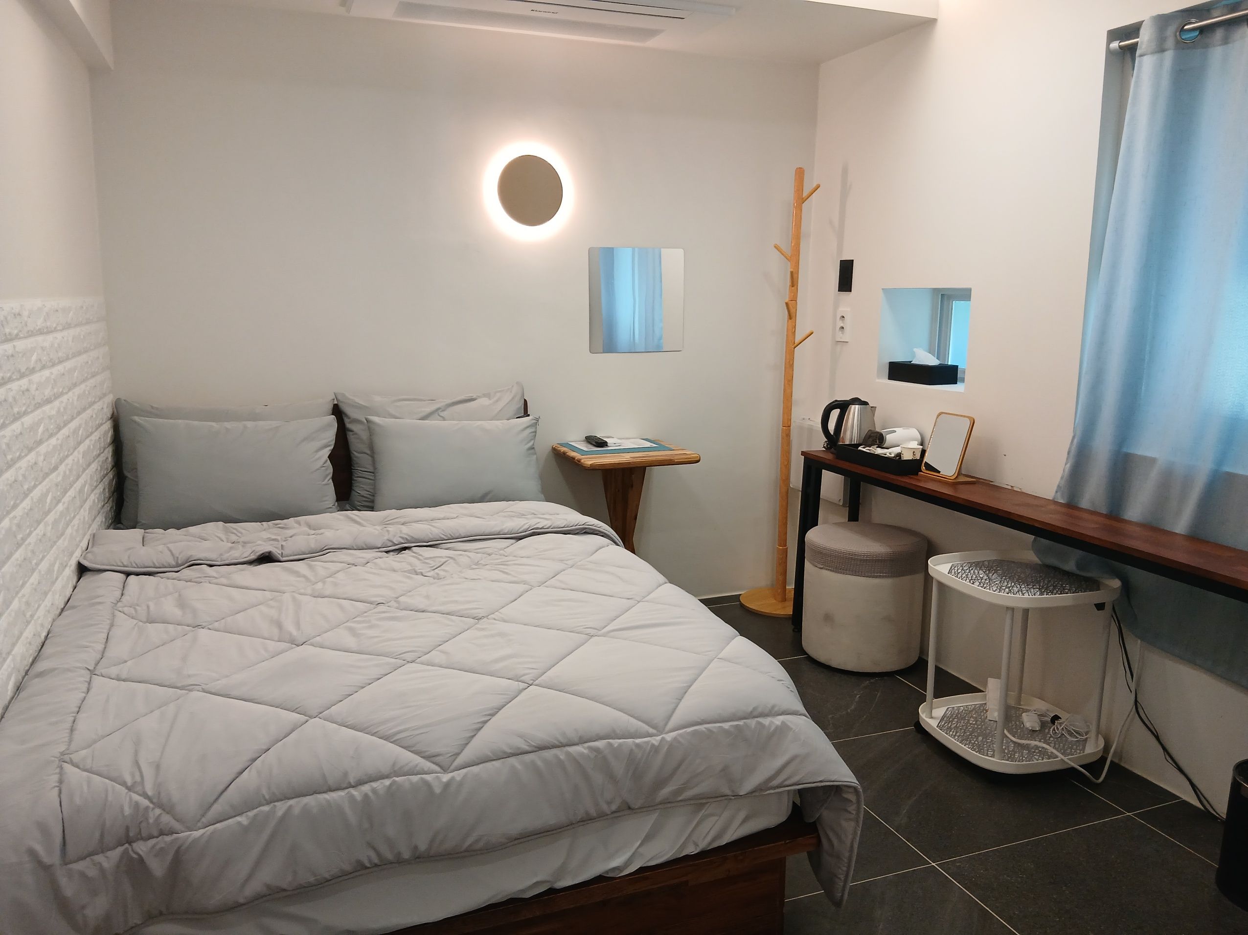 A neatly made bed with a quilted blanket, a bedside table with a kettle and a cup, a small stool, and a trash bin.