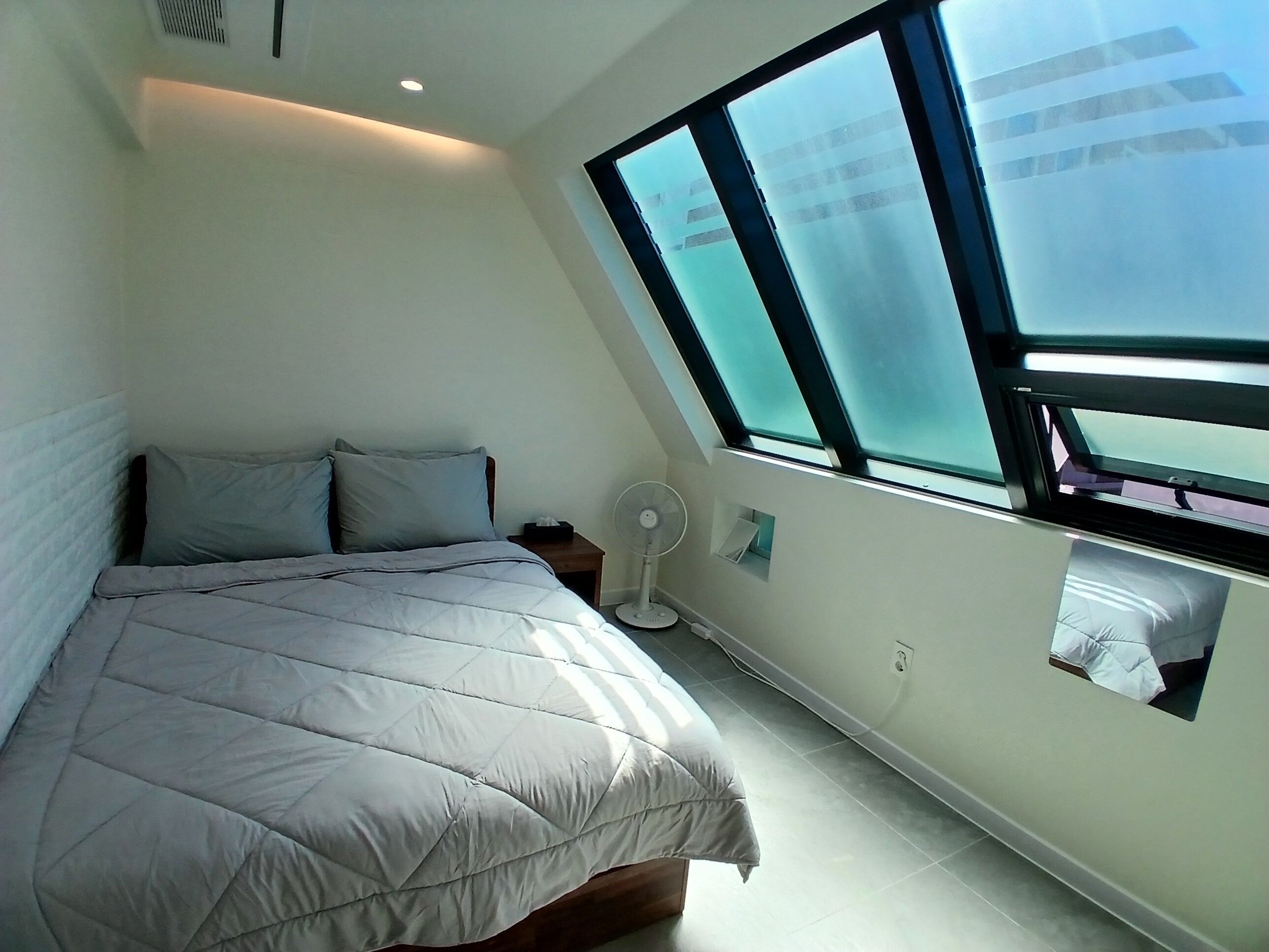 A bedroom with a bed, a nightstand, a wall-mounted fan, and a skylight.