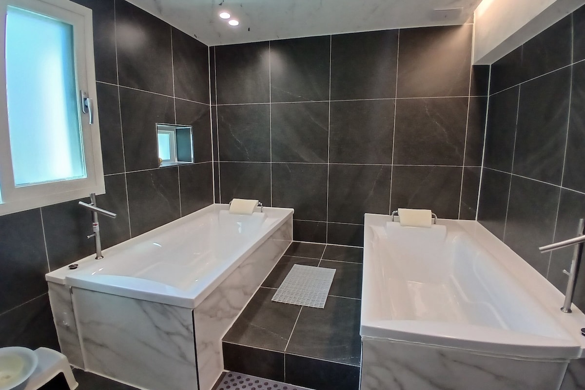 A modern bathroom with two separate tubs, dark tiled walls, and a small window.