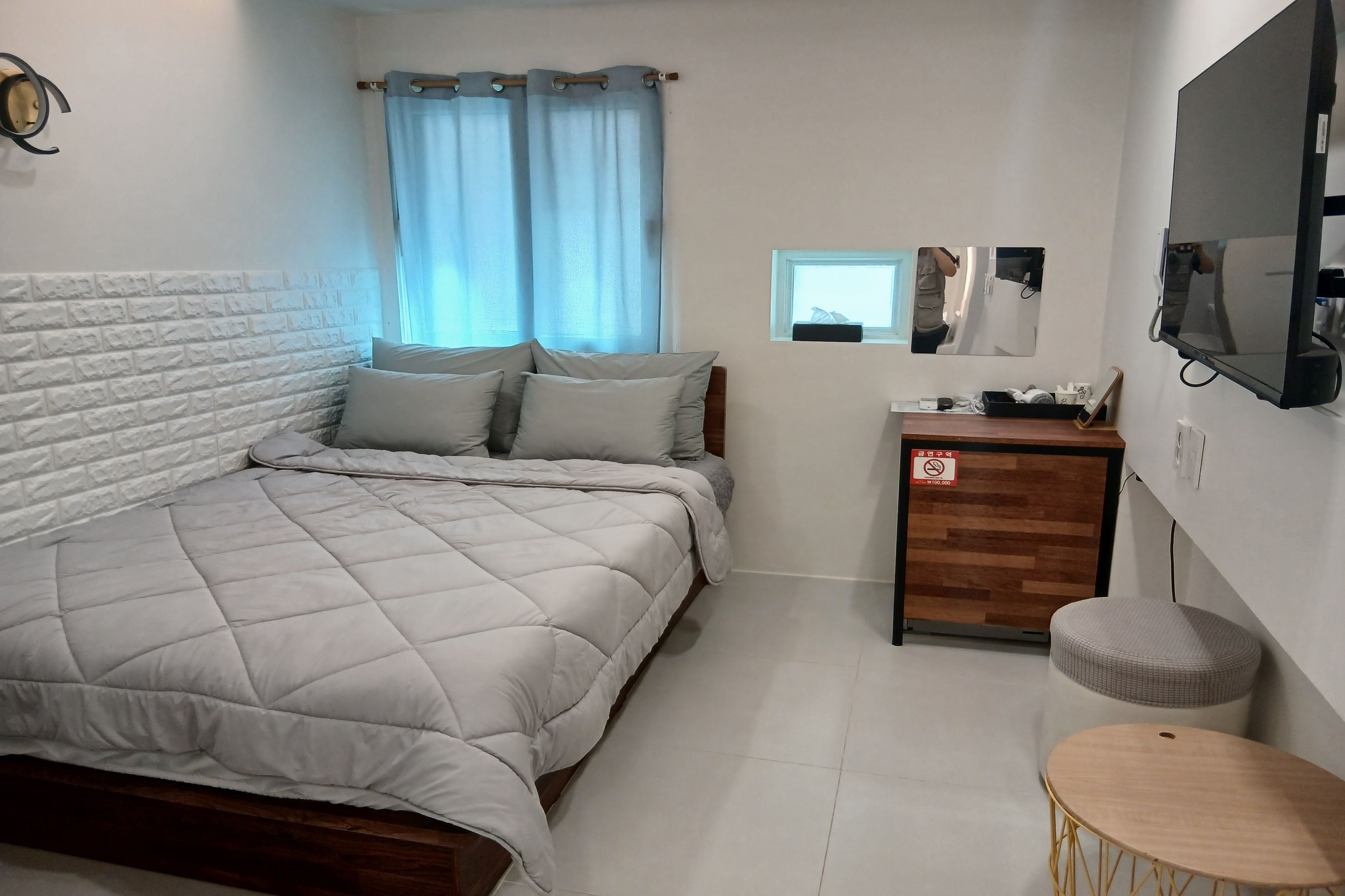 A neatly arranged bedroom with a bed, a small wooden side table, a round stool, and a TV mounted on the wall.