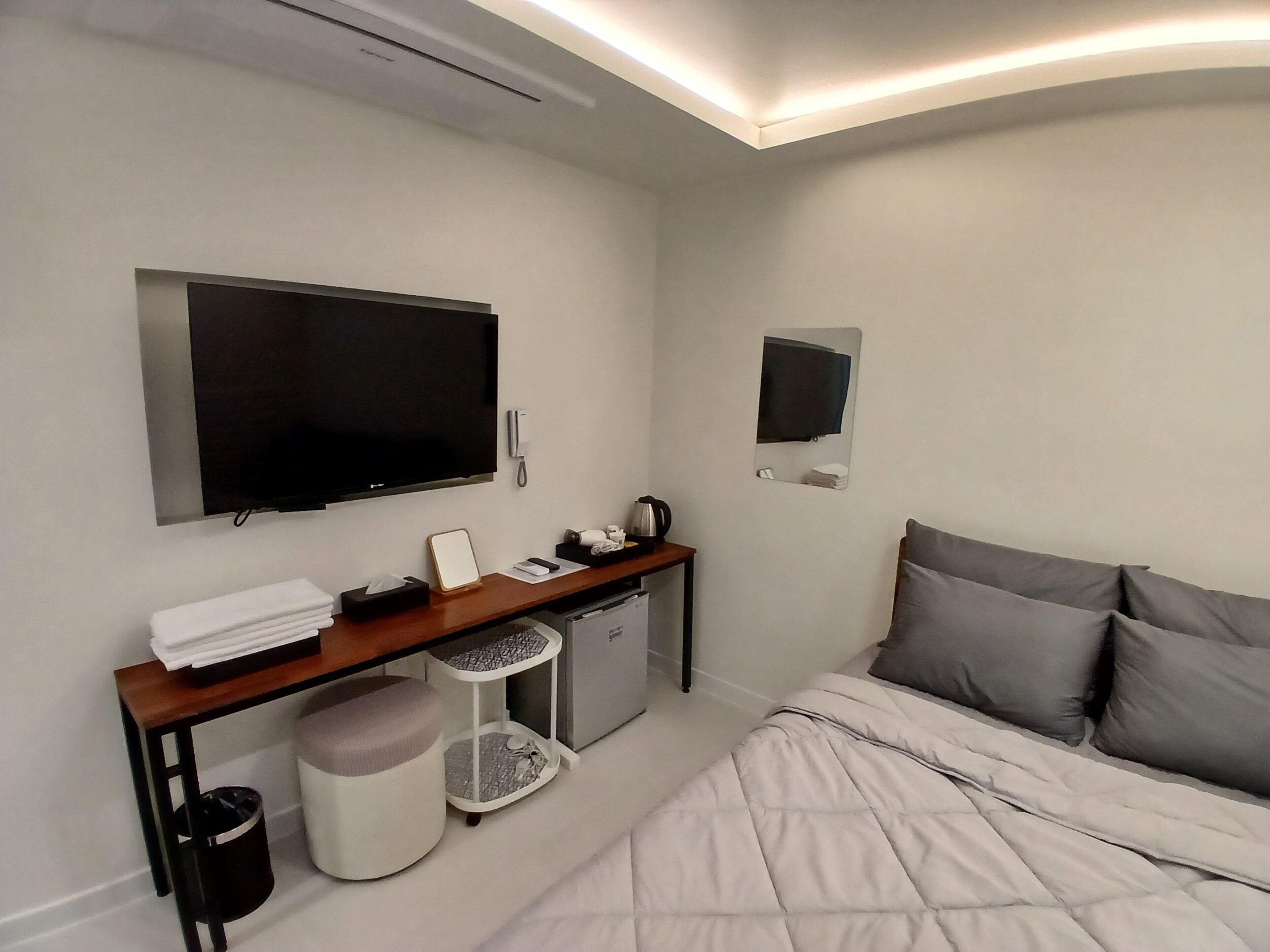 A modern hotel room with a bed, a desk with a flat-screen TV, a small refrigerator, and a trash bin.