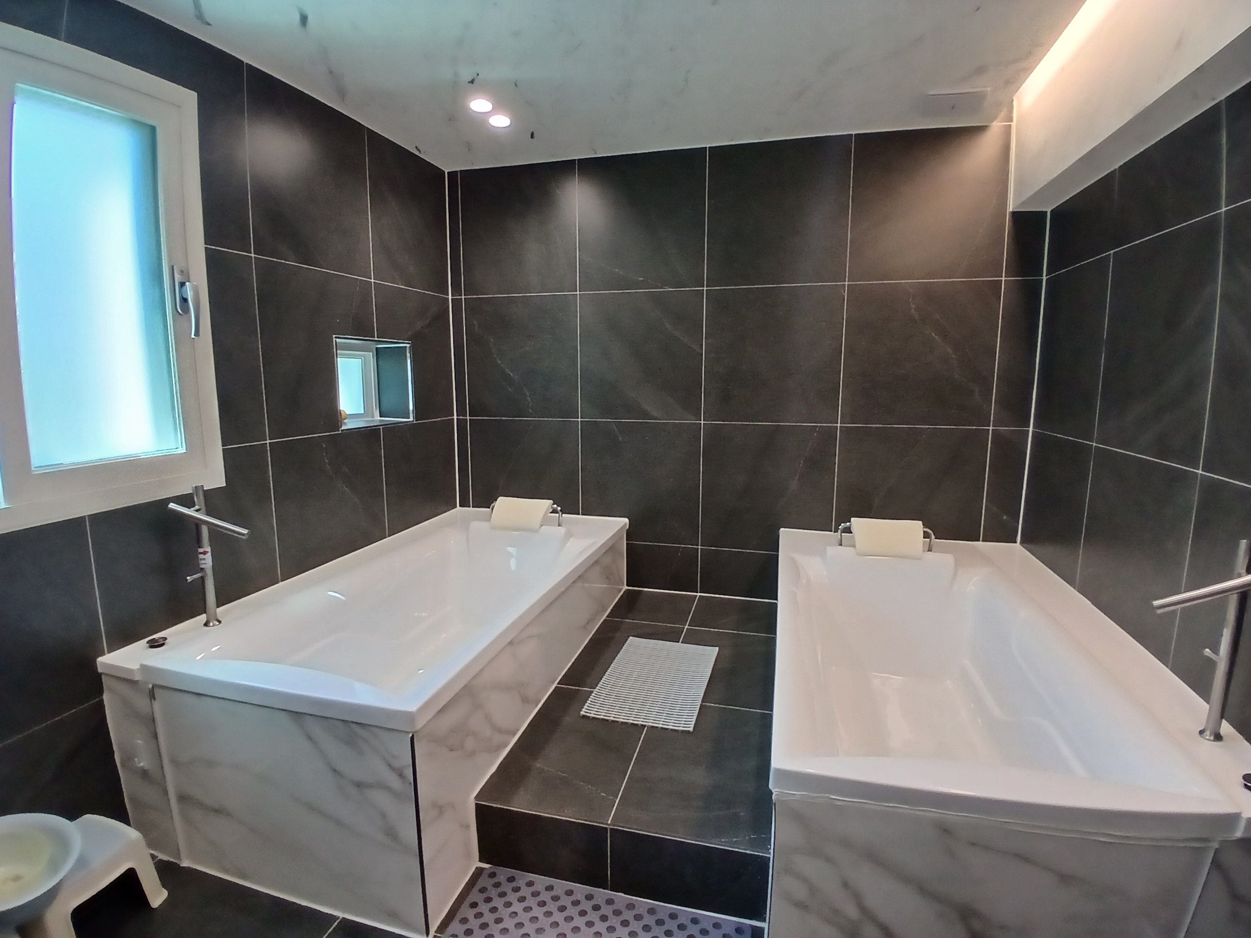 A modern bathroom with two separate tubs, dark tiled walls, and a small window.