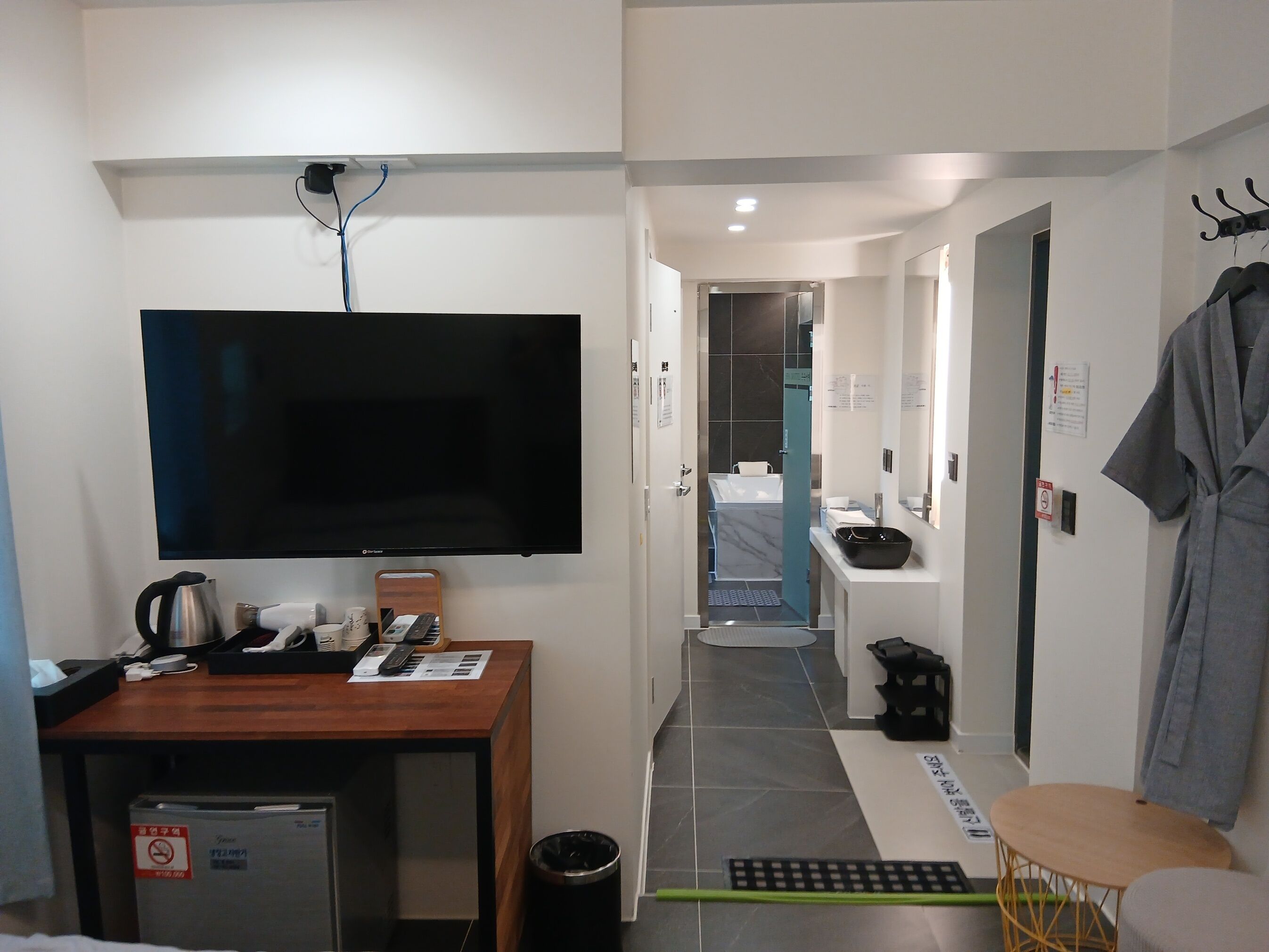 A hotel room with a flat-screen TV, a wooden desk with a kettle, a small fridge, a trash bin, a green mat, a small round table, and a robe hanging on the wall.