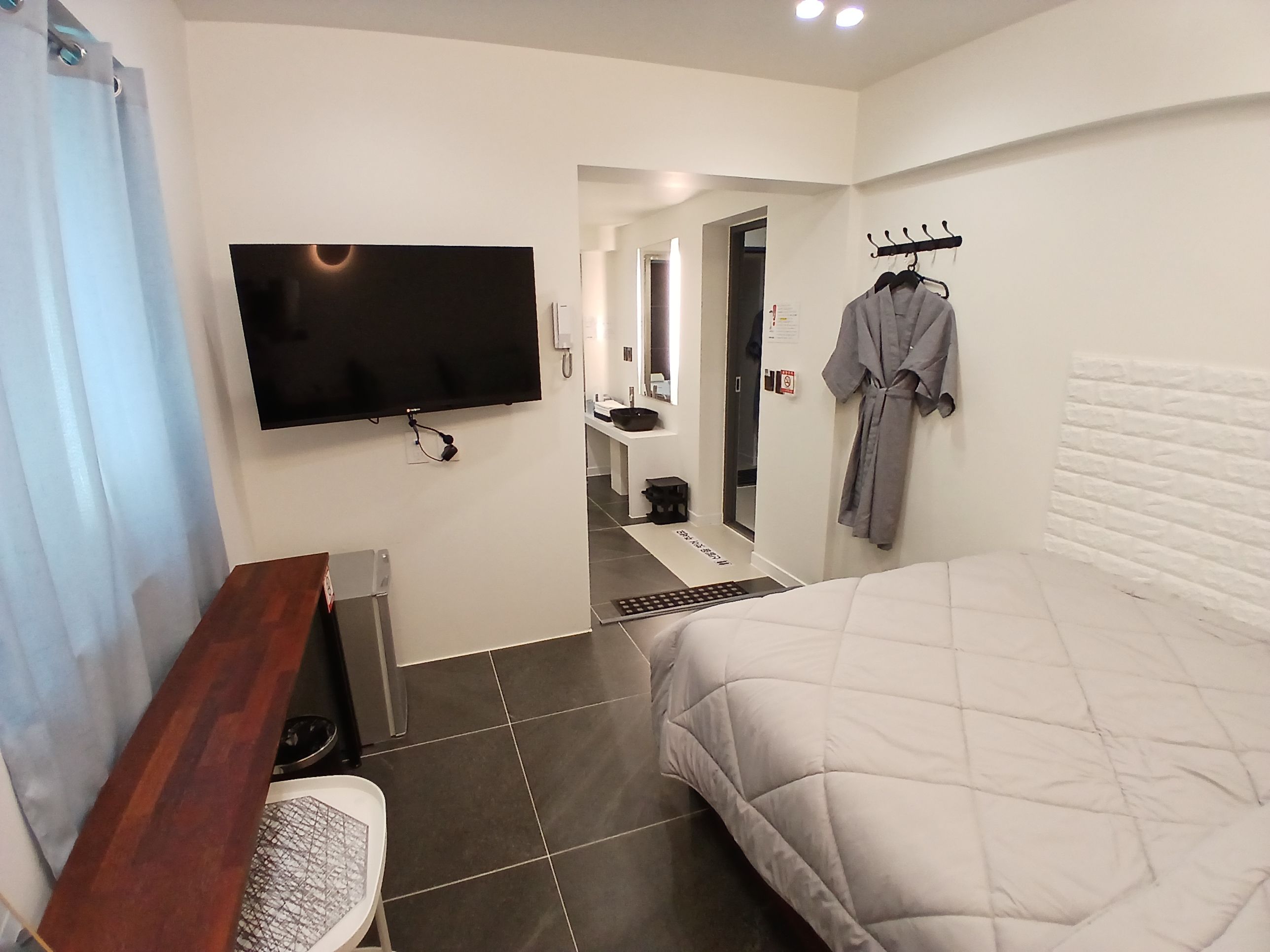 A hotel room with a bed, a TV, a desk, a chair, a robe hanging on the wall, and a bathroom visible through an open door.
