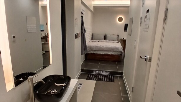 Habitación de hotel moderna con un espejo grande, un lavabo negro, una mesita de noche blanca, una cama con ropa de cama blanca y una alfombra pequeña en el suelo.