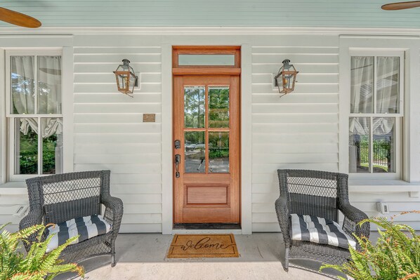 A front porch with a wooden door, two wicker chairs, and a doormat.